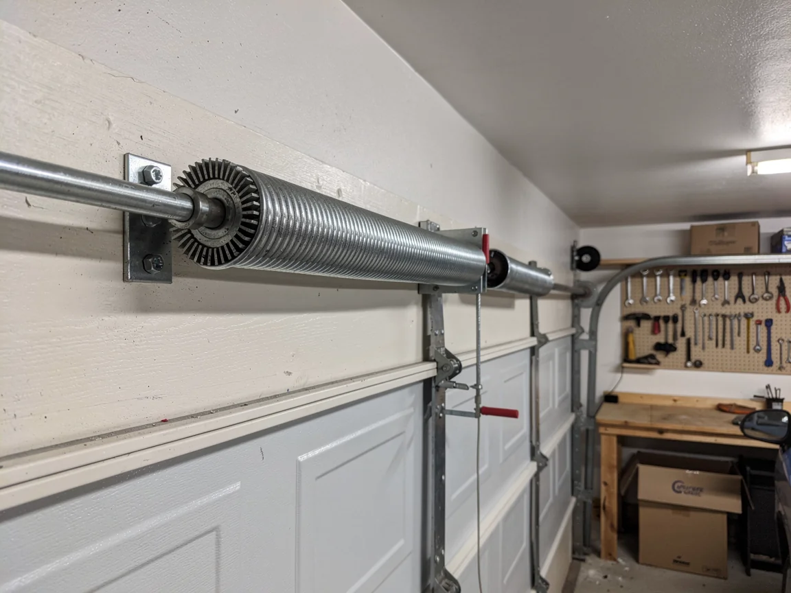 The torsion spring is mounted above a white garage door, with tools visible in the background.